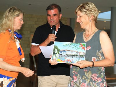 Zwei Frauen stehen bei einer Veranstaltung. Ein Mann spricht und hält ein Bild in der Hand.