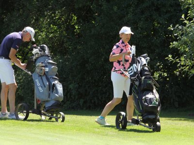 Zwei Personen spielen Golf auf einer Golfanlage. Es ist sonnig und die Umgebung ist grün.