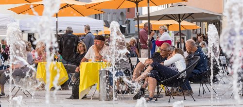 Besucher unterhalten sich angeregt beim Isnyer Feierabendmarkt unter orangenen Sonnenschirmen auf dem Marktplatz.