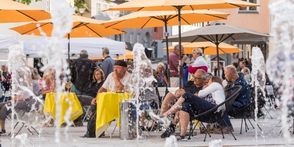 Besucher unterhalten sich angeregt beim Isnyer Feierabendmarkt unter orangenen Sonnenschirmen auf dem Marktplatz.