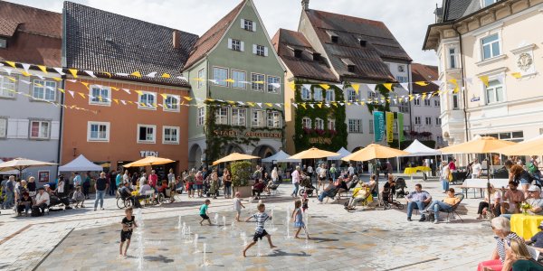 Kinder springen durch das Fontänenfeld auf dem belebten Marktplatz in Isnys Innenstadt.