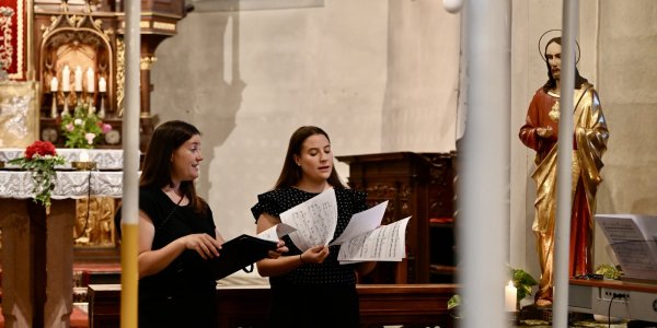Zwei Sängerinnen stehen in einer Kirche und singen während des Gottesdienstes, während eine Statue im Hintergrund steht.