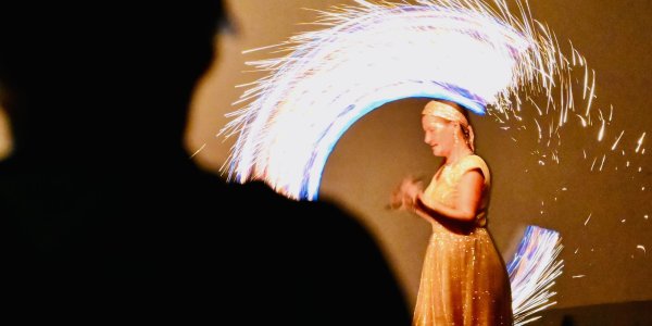 Eine Künstlerin präsentiert eine beeindruckende Feuershow mit hellen Funken im Hintergrund, während Zuschauer gebannt zuschauen.