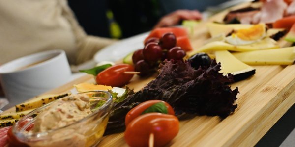 Auf der Holzplatte sind verschiedene Snacks und frische Zutaten angerichtet, während Menschen am Tisch zusammen essen.