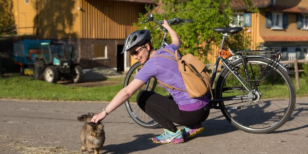Eine Radfahrerin streichelt eine Katze bei einem Bauernhof in Isny