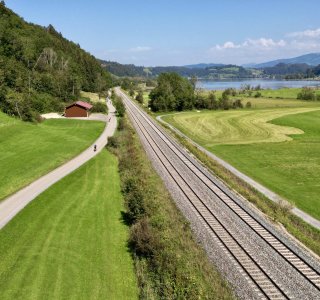 Bahnlinie, daneben eine kleine Straße, im Hintergrund ein See und ein Berg.