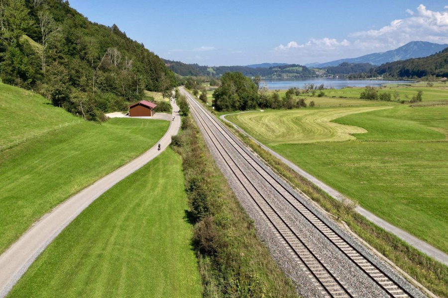 Bahnlinie, daneben eine kleine Straße, im Hintergrund ein See und ein Berg.