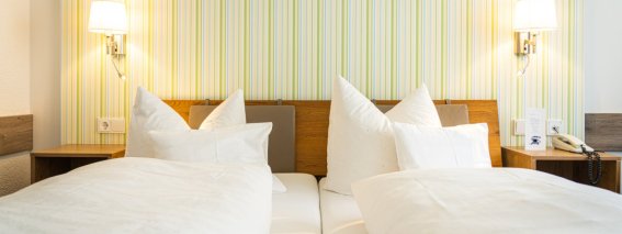 Das Zimmer zeigt zwei Einzelbetten mit weißen Bettdecken und Kissen. Es gibt Lampen und eine einfache Wandgestaltung.