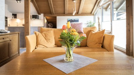 Eine Vase mit bunten Blumen steht auf einem Tisch in einem Wohnzimmer. Im Hintergrund sieht man das Sofa und die Küche.