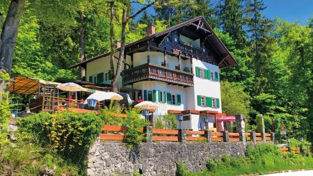 Das Haus steht zwischen Bäumen und hat eine Terrasse. Menschen sitzen draußen und genießen die Natur im Sommer.