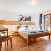 Das Zimmer hat ein großes Bett, einen Tisch und einen Stuhl. Es gibt Fenster, die Licht und Ausblick bieten.