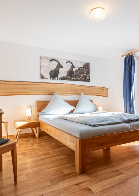 Das Zimmer hat ein großes Bett, einen Tisch und einen Stuhl. Es gibt Fenster, die Licht und Ausblick bieten.