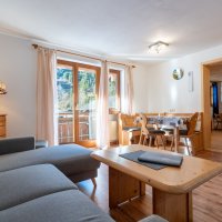 In einem Ferienhaus gibt es einen Wohnraum mit Couch, Tisch und Stühlen. Draußen sind Fenster mit Blick auf die Natur.