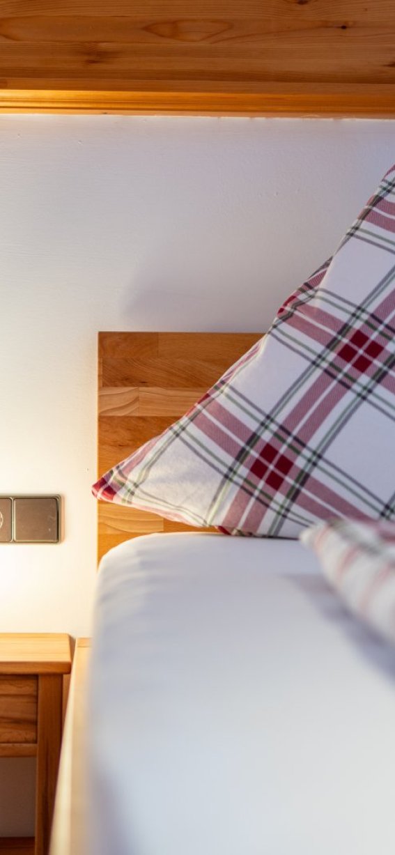 Ein Bett mit kariertem Kissen steht neben einem Nachttisch mit Lampe. Es ist hell beleuchtet und aufgeräumt.