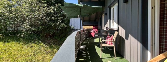 Auf einem Balkon sind zwei Stühle, ein Tisch und ein Sonnenschirm. Der Blick geht auf einen Garten mit grünen Pflanzen.