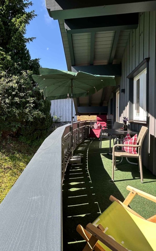Auf einem Balkon sind zwei Stühle, ein Tisch und ein Sonnenschirm. Der Blick geht auf einen Garten mit grünen Pflanzen.