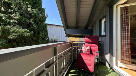 Ein Balkon mit einem roten Stuhl und grünen Pflanzen an einem sonnigen Tag. Die Sonne scheint und es ist windstill.
