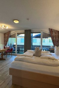 Das Zimmer ist hell und hat einen Balkon. Es gibt einen Tisch, Stühle und ein großes Bett.