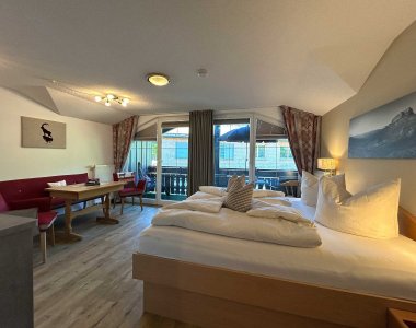 Das Zimmer ist hell und hat einen Balkon. Es gibt einen Tisch, Stühle und ein großes Bett.