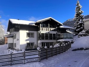 Es liegt Schnee um das Haus in den Bergen. Der Himmel ist blau und die Sonne scheint. Familien besuchen die Gegend.