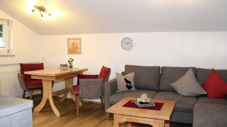 In einem Raum gibt es ein Sofa, einen Tisch und Stühle. Eine Uhr hängt an der Wand. Es ist hell und gemütlich.