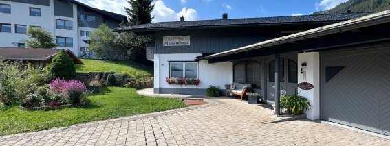 Ein Haus mit einem Garten und gepflasterter Einfahrt. Es steht in einer ruhigen Wohngegend. Wolken sind am Himmel.