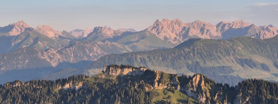 Die Sonne geht über den Bergen auf. Die Landschaft ist grün und hat viele Bäume. Es ist früh am Morgen in den Alpen.