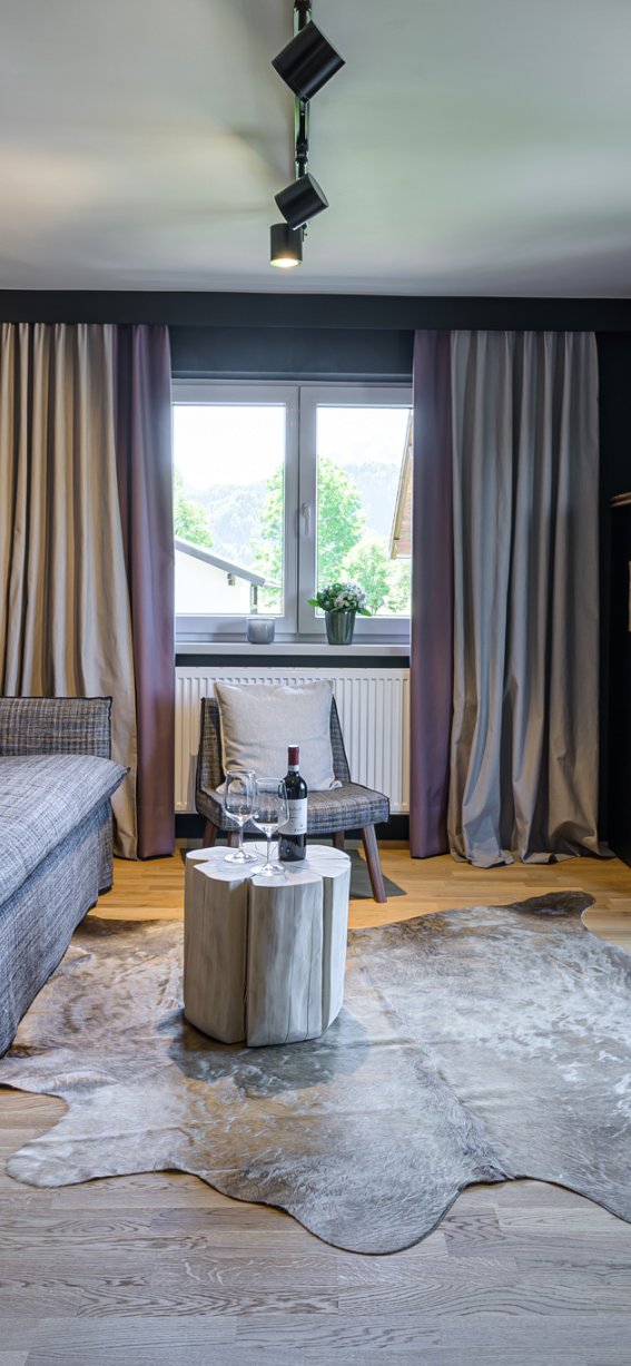 Ein Wohnzimmer mit grauer Couch, Holztisch und Fenstern. Draußen ist es hell und grün. Der Raum hat einen einfachen Stil.