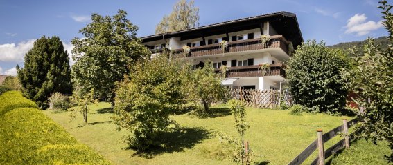 Das Haus steht in einem großen Garten mit Bäumen und Wiesen. Es ist ein sonniger Tag in den Alpen.