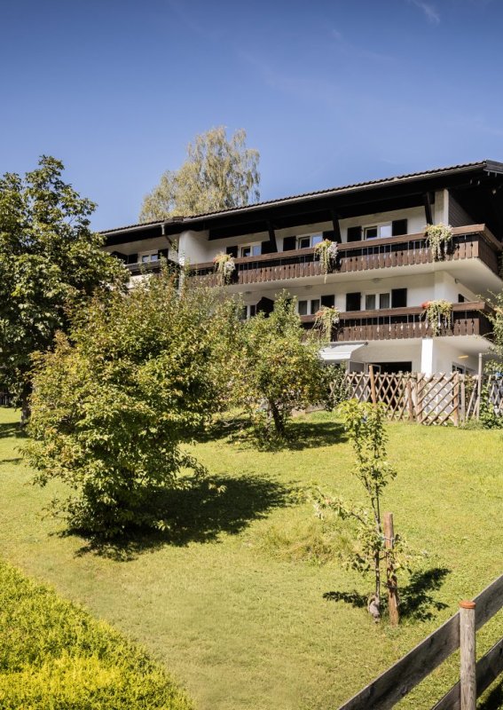 Das Haus steht in einem großen Garten mit Bäumen und Wiesen. Es ist ein sonniger Tag in den Alpen.