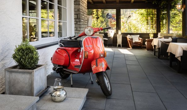 Roter Roller steht vor einem Restaurant im Freien Ein roter Roller steht an einem gediegene Platz vor einem Restaurant. Es ist Tag und die Tische sind bereit.