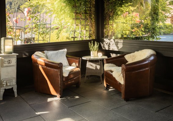Zwei Sessel stehen in einem Gartenpavillon. Pflanzen wachsen um den Raum. Es ist Tag und die Sonne scheint.