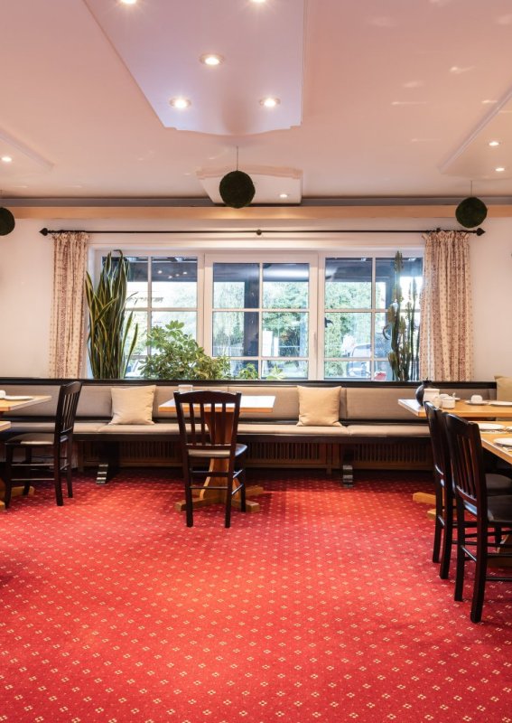Im Restaurant stehen Tische und Bänke. Der Raum hat große Fenster und eine rote Teppichboden.