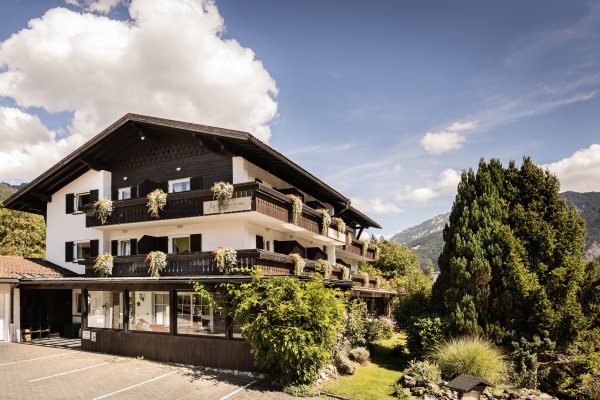 Ein Gebäude im Gebirge mit Balkonen, umgeben von Bäumen und Pflanzen. Es ist ein sonniger Tag mit Wolken am Himmel.