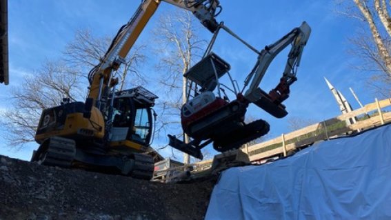 Ein Bagger hebt einen Minibagger auf einer Baustelle im Winter bei klarem Himmel und Bäumen im Hintergrund.