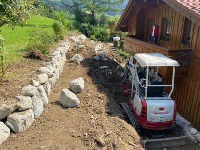 Ein kleiner Bagger bearbeitet den Garten neben einem Holzhaus in den Bergen an einem sonnigen Tag.