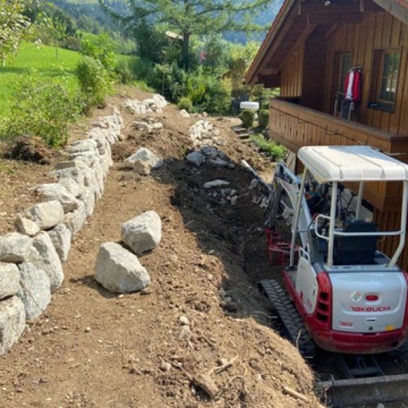 Ein kleiner Bagger bearbeitet den Garten neben einem Holzhaus in den Bergen an einem sonnigen Tag.