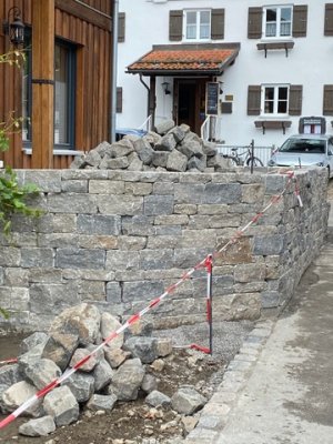 Handwerker arbeiten an einer Steinmauer, die in der Nähe eines Hauses entsteht, umgeben von Bauzubehör und Materialien.