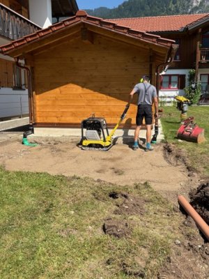 Ein Mann arbeitet im Freien, um den Boden hinter einem Holzhaus zu verdichten und vorzubereiten.