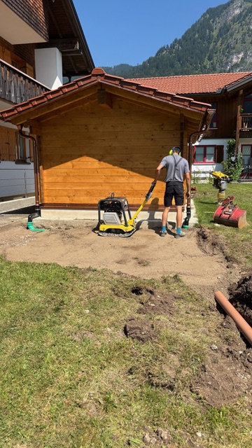 Ein Mann arbeitet im Freien, um den Boden hinter einem Holzhaus zu verdichten und vorzubereiten.