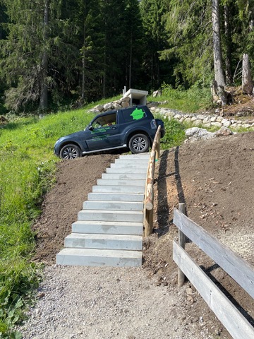 Ein Auto steht ungeschickt auf einer Treppe in einer bergigen Landschaft. Im Hintergrund sind Bäume und eine kleine Hütte sichtbar.
