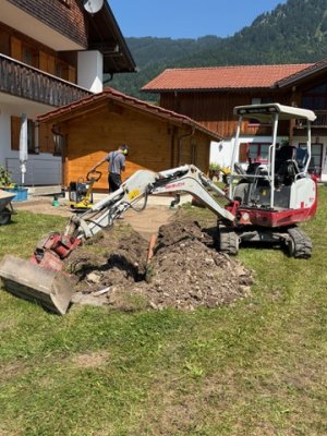 Ein Handwerker mit Mini-Bagger arbeitet im Garten und bewegt Erde, während der Himmel blau und sonnig ist.