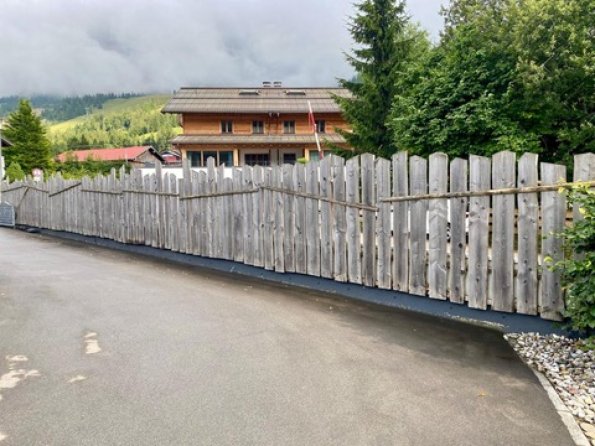 Ein unregelmäßiger Holzlattenzaun umschließt Grundstücke in den bayerischen Alpen. Die Szenerie ist grün und bewaldet.
