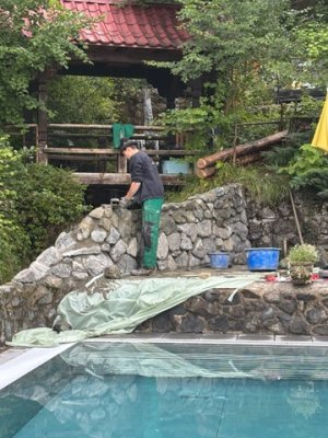 Ein Handwerker verlegt Steine an einer Mauer in einem Gartenbereich neben einem Schwimmbecken.