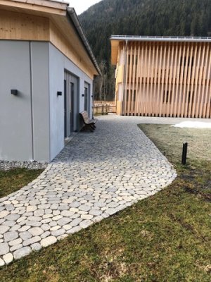 Ein gepflasterter Weg verbindet zwei moderne Gebäude in einer alpinen Umgebung, umgeben von Gras und Bäumen.
