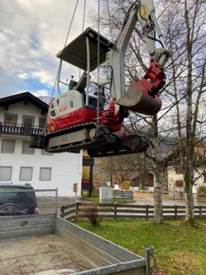 Eine Baustellenmaschine wird von einem Kran über einen Anhänger gehoben, alles findet in einer ländlichen Umgebung statt.