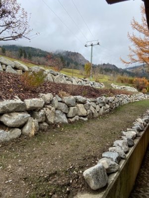 Eine steinerne Mauer flankiert einen Weg in einer herbstlichen Alpenlandschaft mit Wolken und Bergen im Hintergrund.