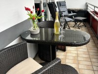 Der Balkon wird von Licht durchflutet. Die Sonne strahlt auf die hellen Fliesen, und die Luft ist frisch und einladend. In der Mitte des Balkons steht ein kleiner, runder Tisch, der perfekt für ein gemütliches Frühstück oder einen entspannten Kaffee am Nachmittag ist. Rund um den Tisch stehen bequeme Klappstühle, die einladend wirken und sich leicht verstauen lassen, wenn du mehr Platz benötigst. Die Stühle sind mit weichen Kissen ausgestattet, die zusätzlichen Komfort bieten und zum Verweilen einladen. Dieser Balkon ist der perfekte Ort, um die warmen Sonnenstrahlen zu genießen, ein gutes Buch zu lesen oder einfach nur die Seele baumeln zu lassen.