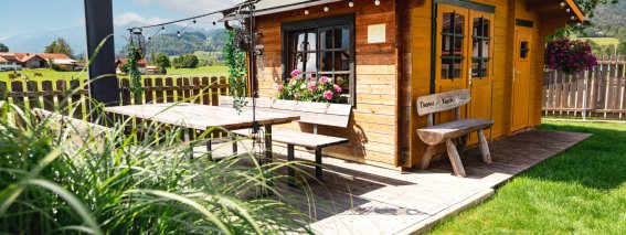 Ein helles Holzhaus steht inmitten einer Wiese mit blühenden Pflanzen und lädt zum Entspannen ein.