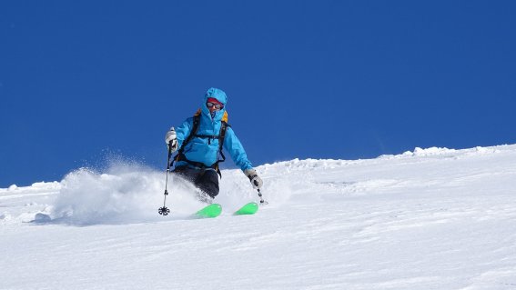 Skifahrer zieht kontrollierte Schwünge durch frischen Tiefschnee unter klarem, blauem Himmel.